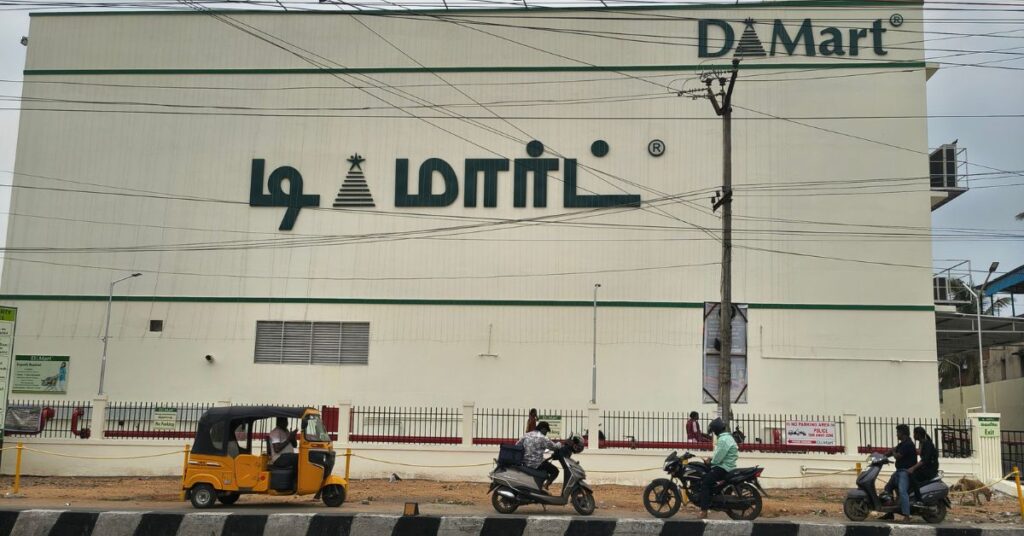 Front view of DMart Avadi building located on Avadi Road in Chennai with Tamil DMart signage, vehicles passing on the road, and the supermarket exterior visible during daytime.